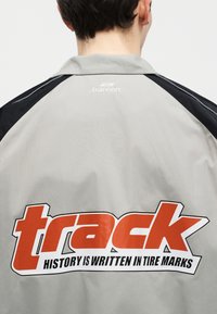 Άτομο που φοράει γκρι και μαύρο μπουφάν με μεγάλο πορτοκαλί κείμενο "track" και μικρότερο σλόγκαν "η ιστορία γράφεται στα σημάδια των ελαστικών" στην πλάτη.