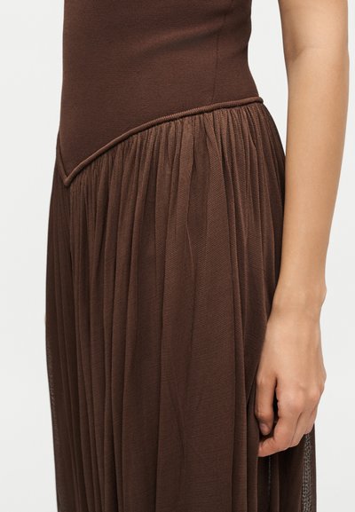 Κοντινό ενός χεριού δίπλα σε καφέ φόρεμα με εφαρμοστό μπούστο και μάζεμένη, πλισέ φούστα.