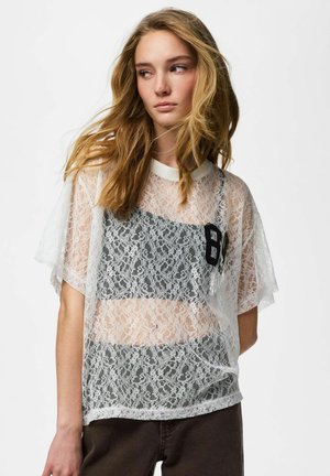 Jeune femme aux longs cheveux blonds portant un haut en dentelle blanche transparente par-dessus un bandeau noir, regardant de côté sur un fond uni.