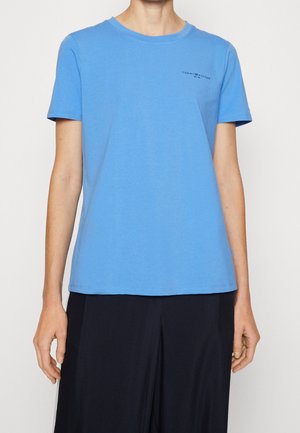 Henkilö pukeutuneena vaaleansiniseen lyhythihaiseen t-paitaan, jossa on pieni "Tommy Hilfiger" -logo vasemmassa rinnassa, yhdistettynä tummiin housuihin.