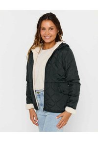 Veste noire matelassée avec une capuche, doublée de fausse fourrure crème. Dispose de poches latérales et d'une fermeture éclair. Portée par-dessus un haut de couleur claire.