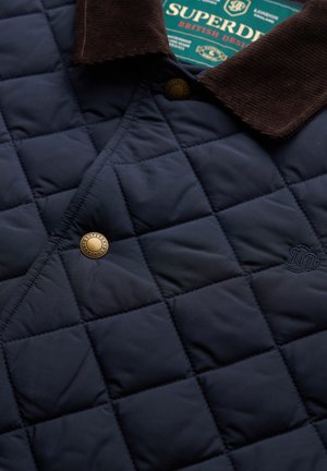 Navy blauw gewatteerde jas met een bruine corduroy kraag, metalen drukknopen en subtiele geborduurde details. Getextureerde, lichte stof.