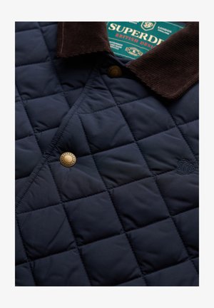 Navy quilted jacket featuring a brown corduroy collar, metal snap closure, and subtle embroidered detail. Textured, lightweight fabric.