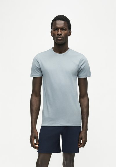 Homme debout portant un t-shirt bleu clair à manches courtes et un short bleu foncé sur un fond blanc uni.