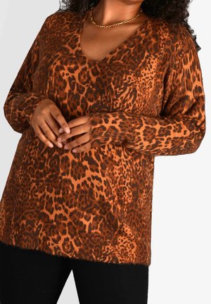 Femme portant un pull à col en V à manches longues avec un motif léopard marron et noir, un pantalon noir et un collier chaîne doré.