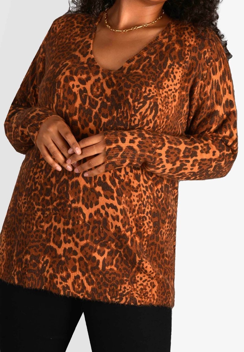 Femme portant un pull à col en V à manches longues avec un motif léopard marron et noir, un pantalon noir et un collier chaîne doré.