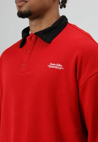 Rode poloshirt met een zwarte kraag, voorzien van vijf knopen en witte geborduurde tekst op de linkerborst. Glad textuur, casual ontwerp.