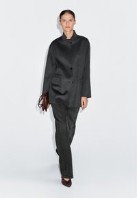 Grå ullblandad kappa med ståkrage, enkel knappstängning och två framfickor, kombinerad med mörka jeans och spetsiga klackar.