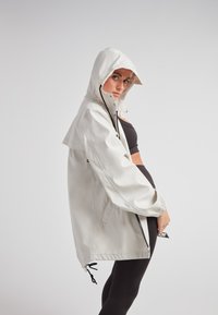 Lichtbeige regenjack met capuchon, voorzien van een ritssluiting aan de voorkant, elastische manchetten en koordjes aan de onderrand voor een verstelbare pasvorm.