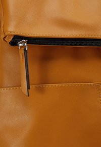 Surface en cuir fauve avec une couture visible et une fermeture éclair noire partiellement ouverte avec une languette en cuir fauve.