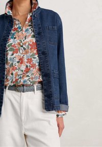 Giacca di jeans in blu scuro, con colletto, chiusura con bottoni e una tasca singola. Sotto, una camicia a stampa floreale con colori vivaci.