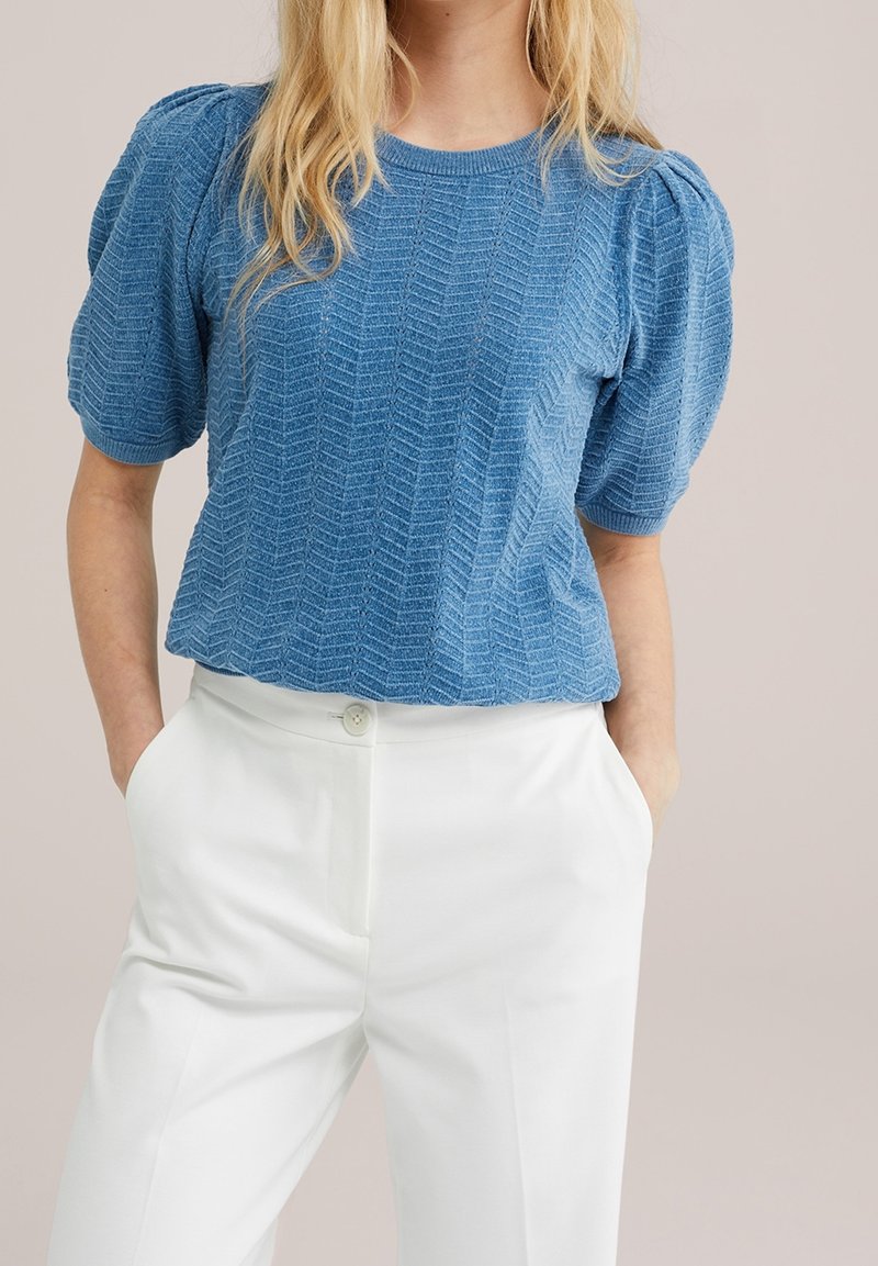 Femme portant un chemisier bleu texturé à manches courtes avec des épaules bouffantes et un pantalon blanc taille haute, les mains dans les poches.