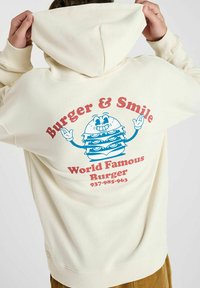 Crème sweatshirt met een grote grafische print op de achterkant van een cartoon hamburger en de tekst "Burger & Smile" in blauw en rood.