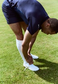 Des chaussures de football blanches avec des détails texturés, portées avec de longues chaussettes blanches, alors qu'un athlète noue ses lacets en se penchant sur l'herbe.