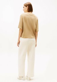 Beiges Kurzarmhemd kombiniert mit weit geschnittenen weißen Hosen, zeigt eine lockere Passform und weiche Baumwollstruktur. Einfaches Design ohne Muster.