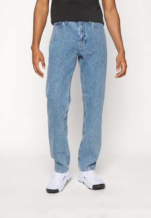 Hombre con vaqueros azul claro, zapatillas blancas y camiseta negra, de pie frente a un fondo claro y liso.