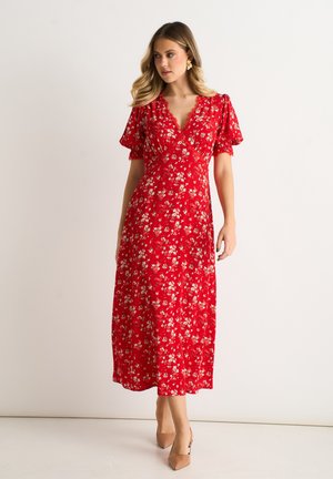 Woman wearing red floral midi dress with puffed short sleeves and V-neck, paired with beige heels, standing against plain background.