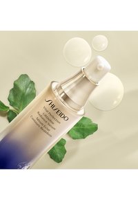 Siero Shiseido in una bottiglia trasparente con un erogatore argentato, sfumatura dal colore oro al blu, accompagnato da gocce e foglie verdi.