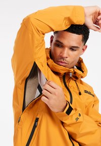 Man die de ventilatierits onder zijn oksel van een feloranje waterdichte jas met capuchon aanpast, tegen een witte achtergrond.