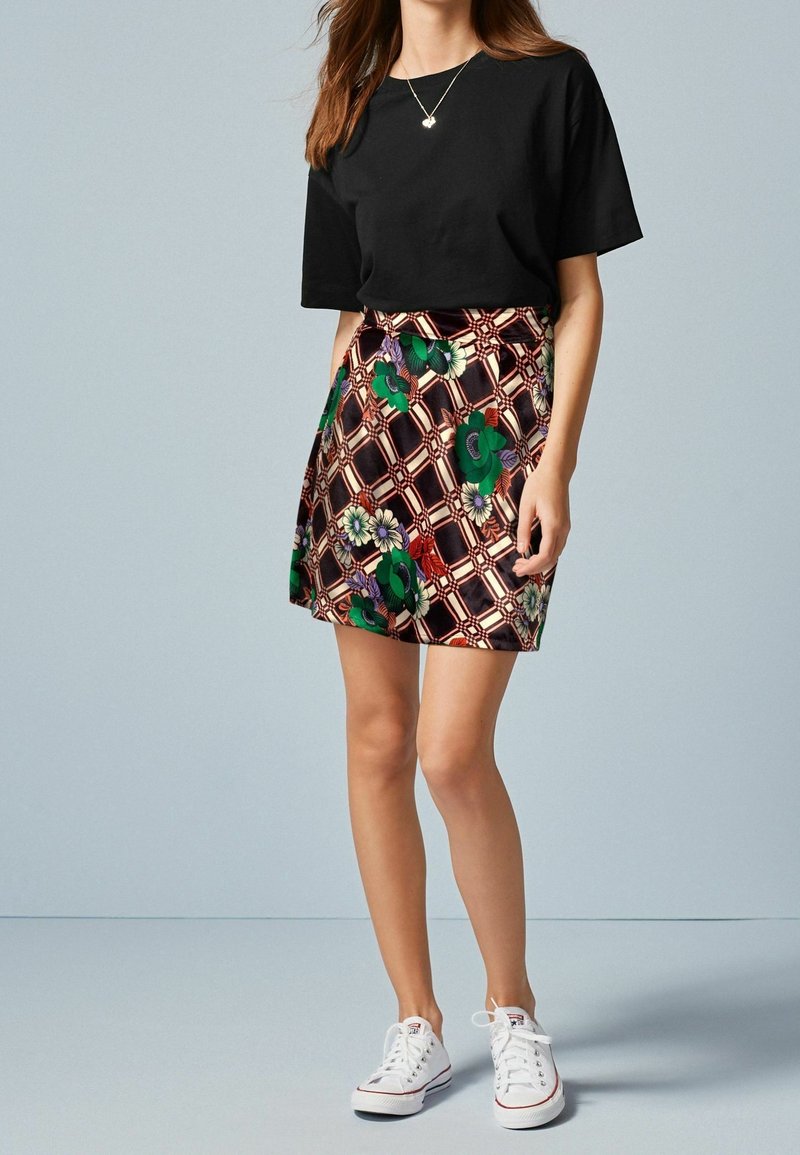 Femme portant un haut noir à manches courtes, une jupe à motif floral avec des fleurs vertes et rouges, et des baskets blanches, debout devant un fond uni.