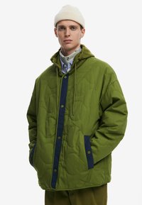 Green quilted jacket with navy accents, snap buttons, a hood, and side pockets. Worn over a striped shirt and a beige beanie.
