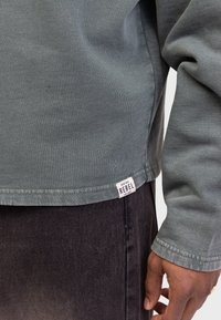 Grijze katoenen sweatshirt met een relaxte pasvorm, zichtbare stiksels en een label met de tekst "REBEL" aan de onderrand, gecombineerd met donkere spijkerbroeken.