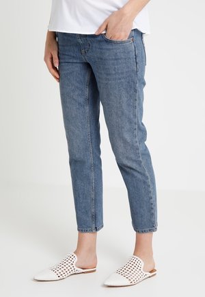 Personne portant un jean bleu et des chaussures blanches en tricot sans lacets, debout avec une main dans la poche sur un fond uni.