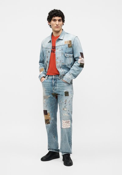 Νεαρός άνδρας που φοράει φωτεινό γαλάζιο τζιν μπουφάν με μπαλώματα και τζιν παντελόνι, κόκκινη μπλούζα με γραφικό σχέδιο και μαύρα αθλητικά παπούτσια, στέκεται με τα χέρια στις τσέπες.