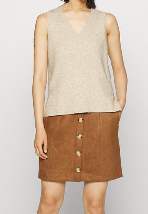 Beige gebreide mouwloze top met een V-hals, gecombineerd met een bruine suède knooprok aan de voorkant. Opvallende variaties in textuur van de materialen.