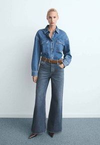 Mujer de pie contra una pared blanca, vistiendo una camisa de mezclilla azul con bolsillos frontales, cinturón marrón, pantalones anchos oscuros y zapatos marrones de punta.