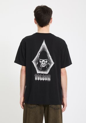 Personne portant un T-shirt noir avec un crâne blanc encapuchonné à l'intérieur d'un design web, avec un texte stylisé en dessous au dos.