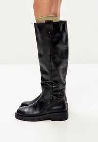 Botas de cuero negro hasta la rodilla con una textura suave, punta redonda, suela resistente y un detalle de puño plegable, complementadas con un calcetín verde oliva.