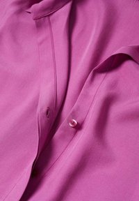 Fuchsia skjorte med glat stof, der har en knaplukning, rund krave og subtile syninger. Knapperne er gennemsigtige.