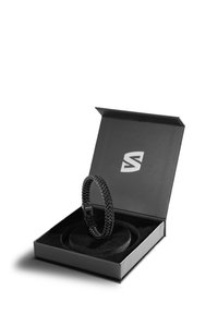 Black woven bracelet with a clasp, displayed in a black box with a soft interior and logo. Box has a smooth finish and slight sheen.
