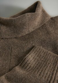 Hnedý pletený vlnený rolák s rebrovaným golierom a manžetami, položený na svetlom povrchu.