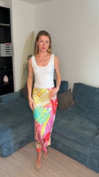 Γυναίκα με μακριά ξανθά μαλλιά που φοράει λευκό τοπ και πολύχρωμη φούστα με σχέδια στέκεται σε σαλόνι με μπλε καναπέ και σανίδα σερφ.