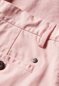 Tela de algodón rosa con detalles de costura, que presenta un botón y un acento metálico etiquetado como "SUPERDRY PREMIUM". Textura suave y forma recta.