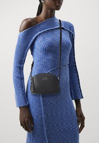 Abito blu a coste con design off-shoulder, caratterizzato da bottoni decorativi; borsa a tracolla nera con texture stampata e dettagli in argento.