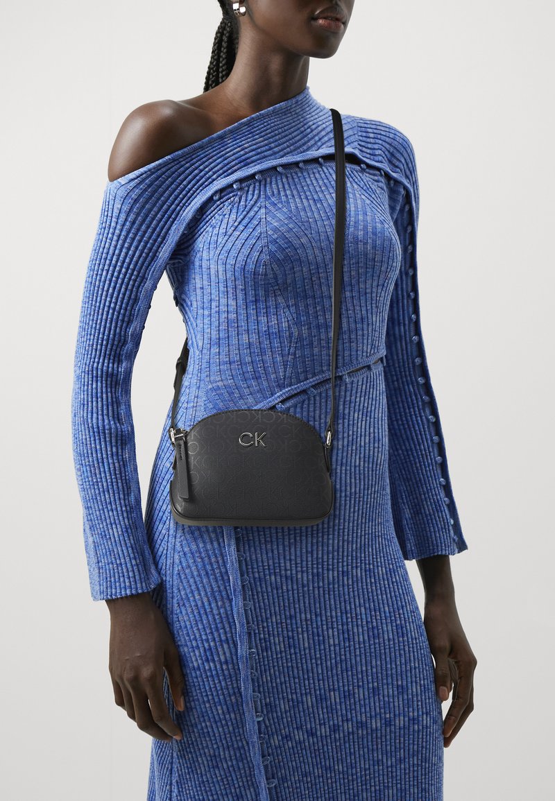 Abito blu a coste con design off-shoulder, caratterizzato da bottoni decorativi; borsa a tracolla nera con texture stampata e dettagli in argento.