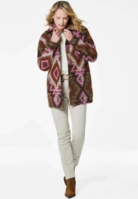 Manteau en laine à motifs marron avec des designs géométriques roses et gris, doté d'un col et de boutons sur le devant, porté sur un pantalon clair.