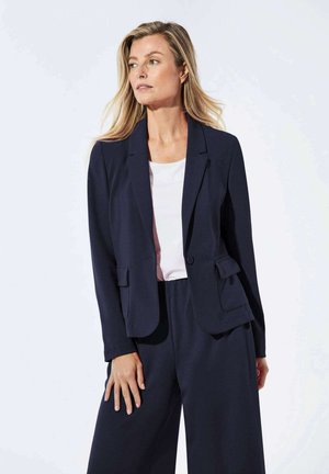 Blazer blu navy dal taglio aderente con chiusura a un bottone, colletto con alette e tasche frontali. Indossato sopra una camicia bianca con pantaloni a gamba larga.