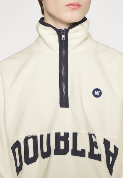 Krémový fleece pulóver s krátkym zipsom a námorníckym zipsom a golierom, s kruhovým námorníckym logom a veľkým textom "DOUBLEA" na spodnej časti.