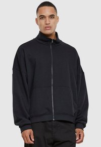 Črna zip-up kapuca z visokim ovratnikom, prevelikim krojem in dolgimi rokavi. Ima gladko teksturo in sprednje žepe.