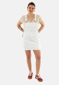 Vestido de tirantes de mezclilla blanca con tirantes ajustables, bolsillos frontales y botones metálicos, combinado con una blusa beige clara y zapatillas rojas.