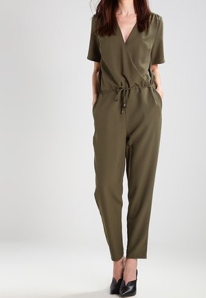 Kobieta w oliwkowozielonym, krótkim kombinezonie kopertowym z paskiem w talii i czarnych szpilkach ze szpiczastym noskiem, stojąca z rękami w kieszeniach.