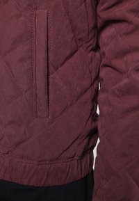 Giacca trapuntata di un profondo colore bordeaux, con tasca laterale, orlo arricciato e texture liscia caratterizzata da cuciture a rombi evidenti.