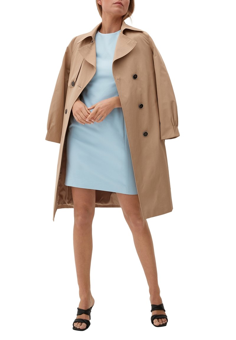 Manteau beige en trench avec un design croisé, grands revers et boutons noirs, porté sur une robe bleu clair, associé à des sandales noires à brides.