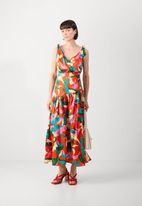 Robe sans manches colorée avec un décolleté en V, jupe froncée, motifs floraux en rouge, rose, vert et orange. Assortie à des sandales à talons rouges.