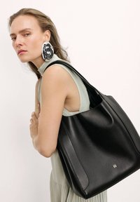 Czarna skórzana torba tote o gładkiej teksturze i miękkim, ustrukturyzowanym kształcie. Posiada złote okucia z logo oraz szeroki pasek do noszenia na ramieniu.