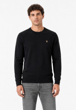 Uomo con capelli ricci che indossa un maglione nero a maniche lunghe e jeans grigi che sta in piedi davanti a uno sfondo bianco uniforme.